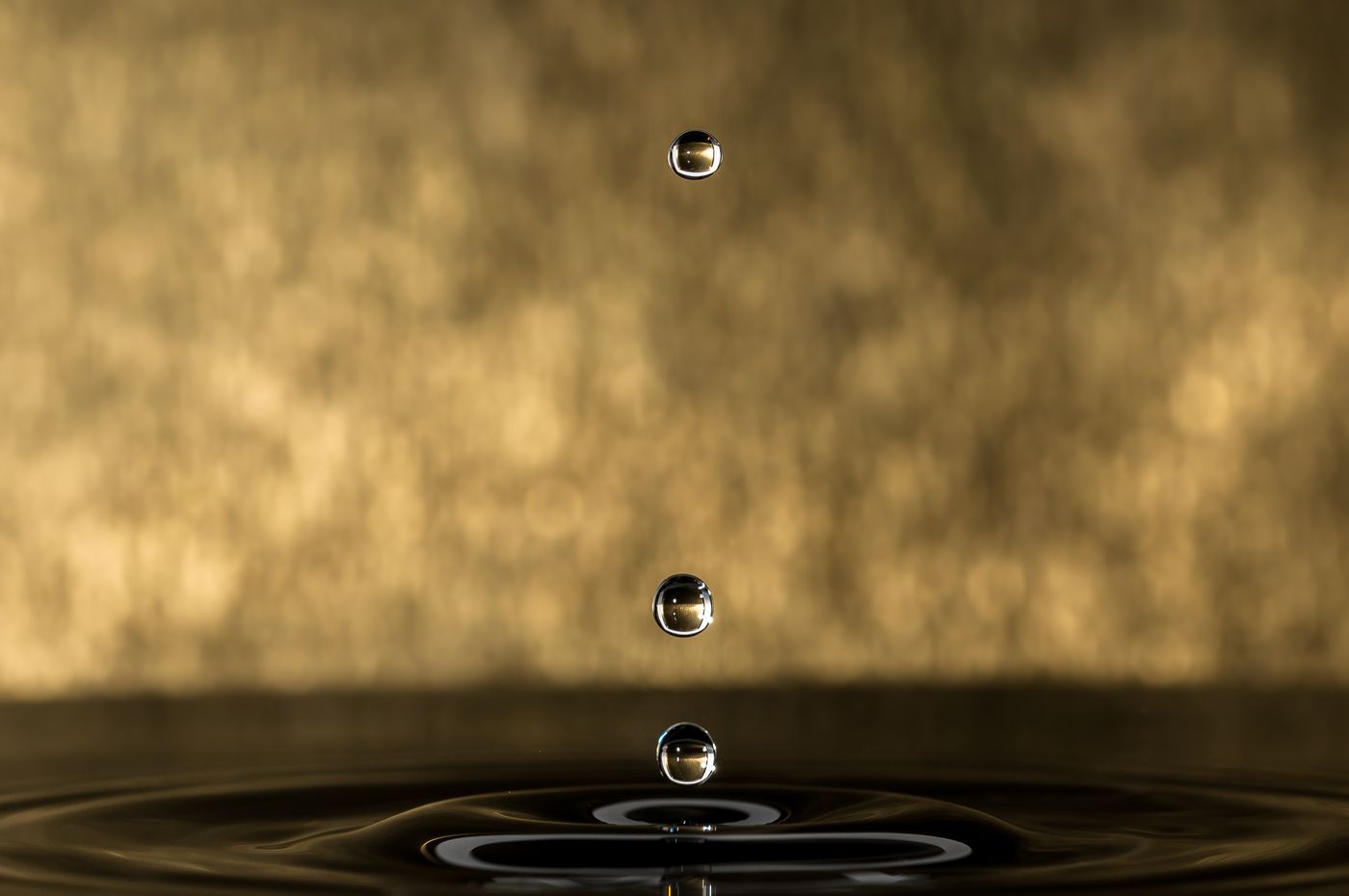 Drei fallende Öltropfen über einer Wasseroberfläche vor goldenem Hintergrund